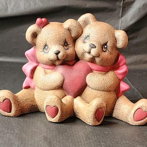 Könnte beinhalten: Zwei braune Teddybären, die ein rotes Herz halten. Die Bären tragen rosa Schleifen und sitzen nebeneinander.