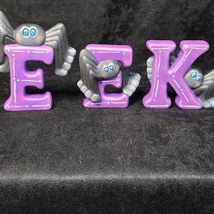 May include: Three purple letters spelling the word EEK, each featuring a grey spider with blue eyes attached to the surface. The letters have white painted accents and dots along their edges, creating a decorative set of freestanding alphabet characters.