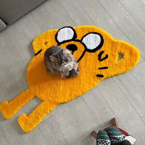 Puede incluir: Una alfombra amarilla y negra con forma de perro de dibujos animados con un gato gris sentado encima. La alfombra tiene un dise&ntilde;o de ojos blanco y negro.