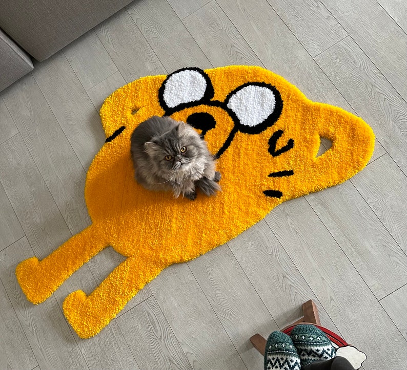 Puede incluir: Una alfombra amarilla y negra con forma de perro de dibujos animados con un gato gris sentado encima. La alfombra tiene un dise&ntilde;o de ojos blanco y negro.