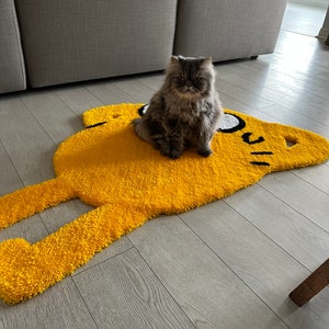 Puede incluir: Un gato gris sentado en una alfombra amarilla con forma de personaje de dibujos animados. La alfombra tiene contornos negros y ojos blancos.