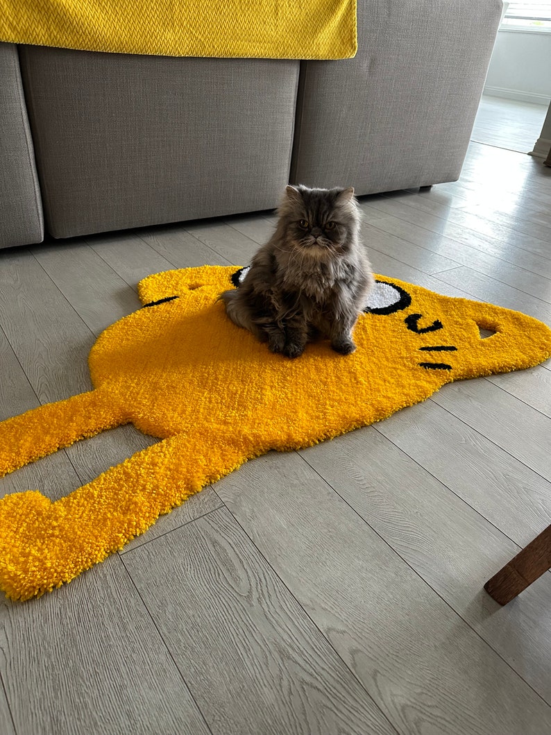Puede incluir: Un gato gris sentado en una alfombra amarilla con forma de personaje de dibujos animados. La alfombra tiene contornos negros y ojos blancos.