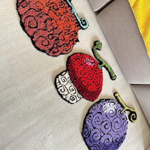May include: Three tufted rugs shaped like stylized fruits with swirling patterns. One rug is orange and red with a light blue stem, one is red and white with a green stem, and one is purple and black with a green stem, all featuring black outlines.