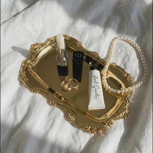 Puede incluir: Una bandeja dorada y ornamentada contiene productos de belleza y joyas. Los artículos incluyen un lápiz labial, un tubo negro, un pequeño tubo de crema de manos con el texto "Aunt Gentiic", dos anillos dorados y un collar de perlas. La bandeja está sobre una superficie blanca.
