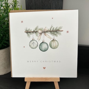 May include: A Christmas card with a watercolour illustration. A sprig of pine branches with a bow and three hanging baubles in shades of green and white. The words "MERRY CHRISTMAS" are printed below the baubles.