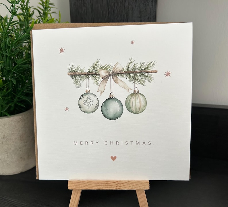 May include: A Christmas card with a watercolour illustration. A sprig of pine branches with a bow and three hanging baubles in shades of green and white. The words "MERRY CHRISTMAS" are printed below the baubles.