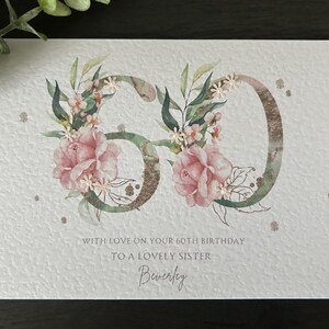 Puede incluir: Tarjeta de cumplea&ntilde;os para el 60 cumplea&ntilde;os, con el n&uacute;mero 60 adornado con flores rosas y hojas verdes. La tarjeta incluye el texto "Con amor en tu 60 cumplea&ntilde;os a una querida hermana" y el nombre "Beverley".