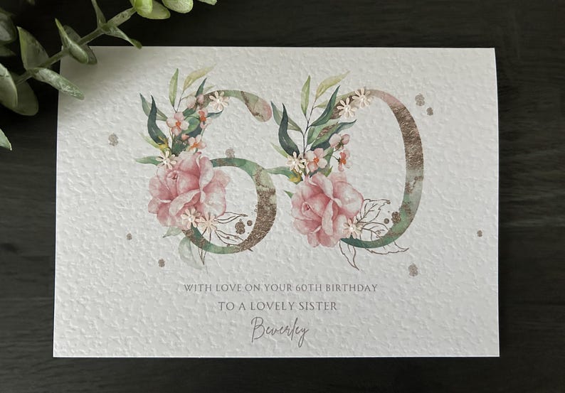 Puede incluir: Tarjeta de cumplea&ntilde;os para el 60 cumplea&ntilde;os, con el n&uacute;mero 60 adornado con flores rosas y hojas verdes. La tarjeta incluye el texto "Con amor en tu 60 cumplea&ntilde;os a una querida hermana" y el nombre "Beverley".