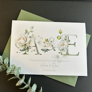 May include: A wedding card featuring the initials "A & E" adorned with floral designs. The card rests on a green envelope, with the text "Congratulations on your wedding day", names "Anne & Eric" and the date "17th August 2025".