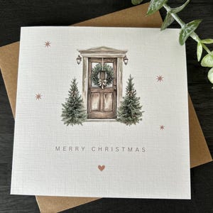 May include: A square Christmas card featuring a watercolour illustration of a wooden door decorated with a wreath, with two small Christmas trees on either side. The words "MERRY CHRISTMAS" are printed below, with a heart symbol.