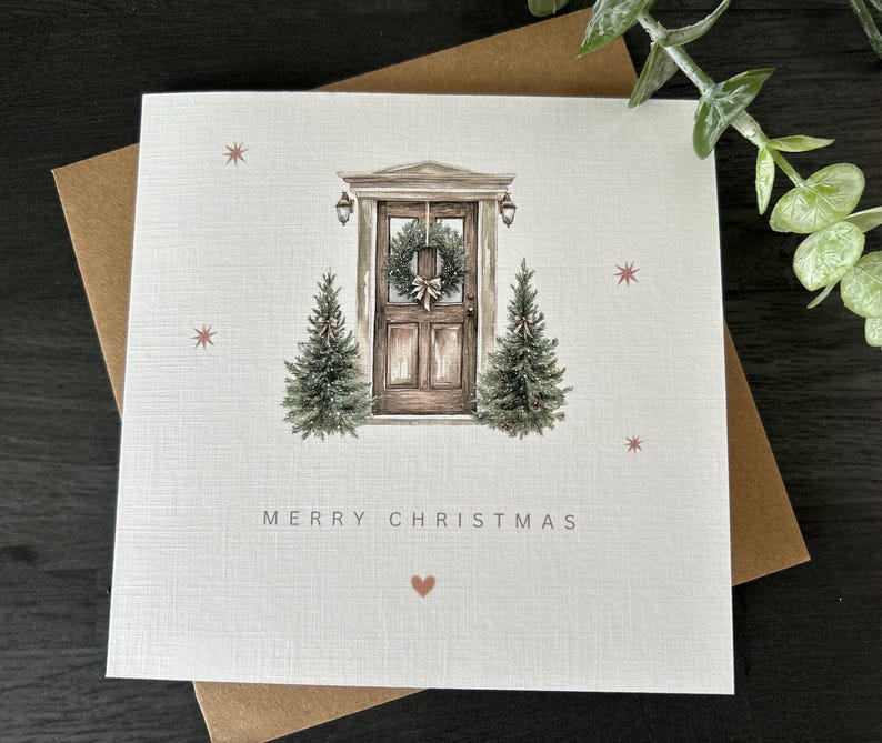 May include: A square Christmas card featuring a watercolour illustration of a wooden door decorated with a wreath, with two small Christmas trees on either side. The words "MERRY CHRISTMAS" are printed below, with a heart symbol.