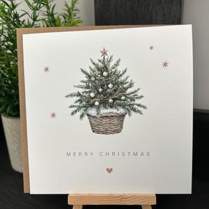 May include: A square Christmas card featuring a watercolour illustration of a decorated Christmas tree in a woven basket. The card reads "MERRY CHRISTMAS" with a heart symbol. Small star and snowflake accents are scattered around the tree.