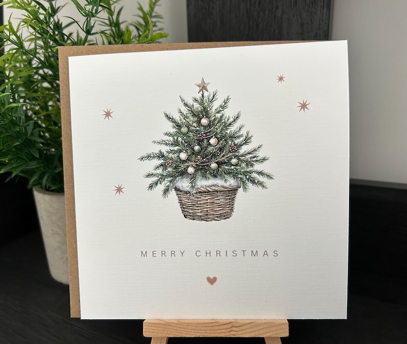 May include: A square Christmas card featuring a watercolour illustration of a decorated Christmas tree in a woven basket. The card reads "MERRY CHRISTMAS" with a heart symbol. Small star and snowflake accents are scattered around the tree.