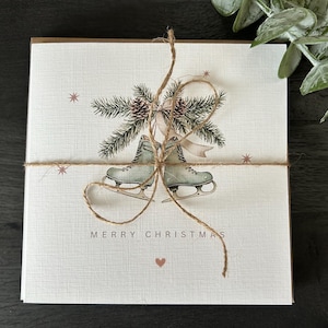May include: A stack of Christmas cards tied with twine. Each card features a watercolour illustration of ice skates, pine boughs, and the words "MERRY CHRISTMAS". The cards have a textured, off-white background with small star and heart accents.