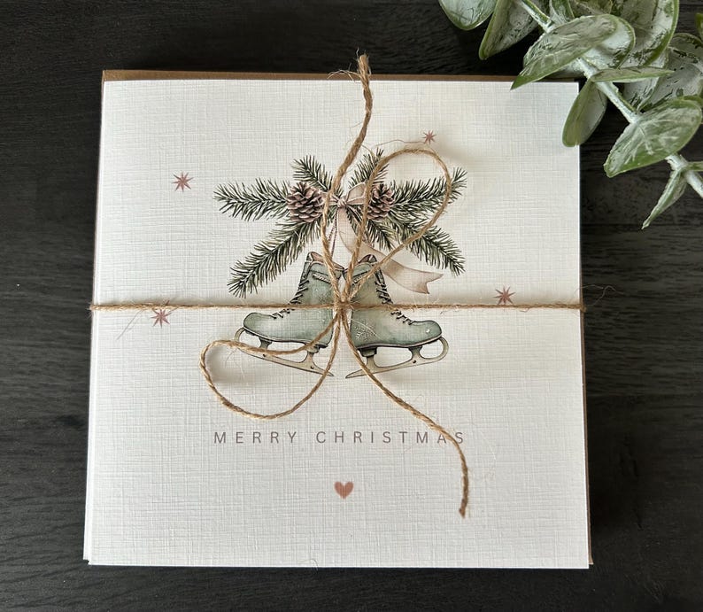 May include: A stack of Christmas cards tied with twine. Each card features a watercolour illustration of ice skates, pine boughs, and the words "MERRY CHRISTMAS". The cards have a textured, off-white background with small star and heart accents.