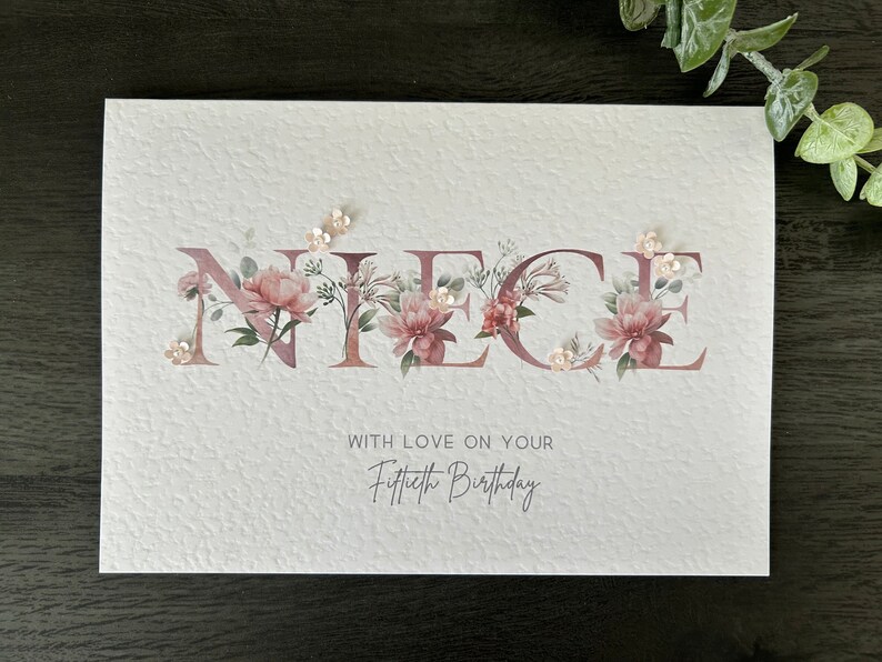 K&ouml;nnte beinhalten: Eine wei&szlig;e Karte mit dem Wort "NIECE" in pinken Blumenbuchstaben. Die Karte tr&auml;gt den Text "With love on your Fiftieth Birthday" unter den Blumenbuchstaben.