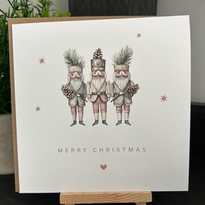 May include: A Christmas card with a watercolour illustration of three nutcracker soldiers. The card has the words "MERRY CHRISTMAS" and a small heart. The nutcrackers are in green, pink, and beige, holding pine cones. The card is on a wooden easel.