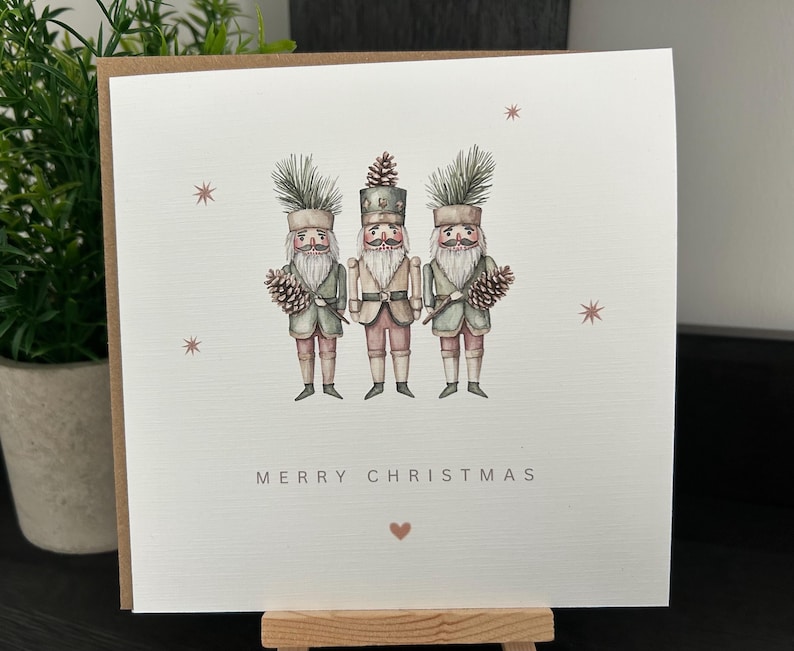 May include: A Christmas card with a watercolour illustration of three nutcracker soldiers. The card has the words "MERRY CHRISTMAS" and a small heart. The nutcrackers are in green, pink, and beige, holding pine cones. The card is on a wooden easel.