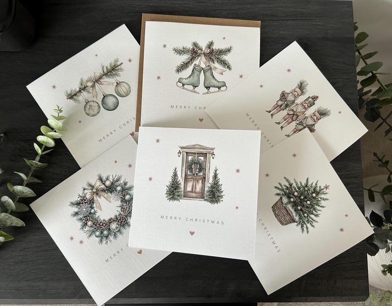 May include: A selection of Christmas cards featuring watercolour illustrations. Designs include ice skates, baubles, a wreath, a door, and a basket of greenery. Each card has the words "MERRY CHRISTMAS" printed on it.