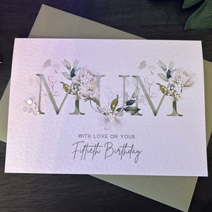 May include: A greeting card with the word "MUM" cut out, decorated with floral accents. The card reads "WITH LOVE ON YOUR Fiftieth Birthday" in elegant script. The card is on a green envelope.