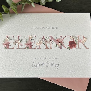 May include: A personalized birthday card with the name "Eleanor" in mauve letters, adorned with floral arrangements. The card reads "To a special friend" and "With love on your Eightieth Birthday."