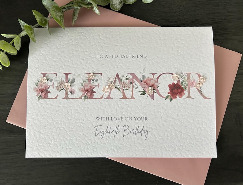 May include: A personalized birthday card with the name "Eleanor" in mauve letters, adorned with floral arrangements. The card reads "To a special friend" and "With love on your Eightieth Birthday."