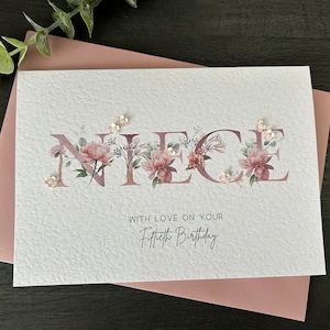 K&ouml;nnte beinhalten: Eine wei&szlig;e Karte mit dem Wort "NIECE" in rosa Buchstaben mit floralen Designs. Die Karte tr&auml;gt den Text "With love on your Fiftieth Birthday" unter dem Wort "NIECE".