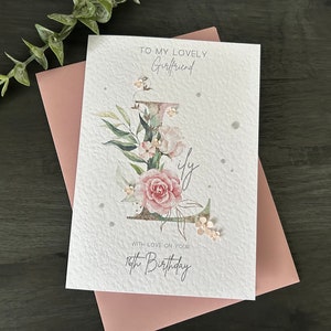 May include: A white card with a pink floral design and the text "To my lovely Girlfriend" and "With love on your 18th Birthday".
