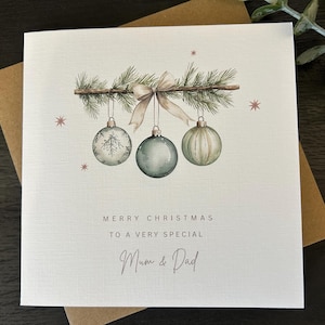 May include: A Christmas card with a watercolour design. The card features three hanging baubles in shades of green and white, suspended from a branch with a bow. The text reads "Merry Christmas to a very special Mum & Dad."