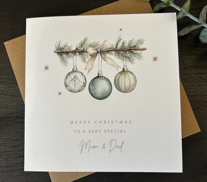 May include: A Christmas card with a watercolour design. The card features three hanging baubles in shades of green and white, suspended from a branch with a bow. The text reads "Merry Christmas to a very special Mum & Dad."
