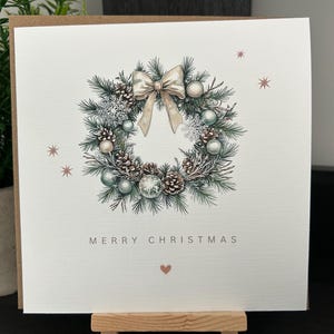 May include: A Christmas card with a watercolour illustration of a wreath. The wreath is composed of pine needles, pine cones, and baubles in green, white, and silver. A bow sits at the top, and the words "MERRY CHRISTMAS" are printed below the wreath.