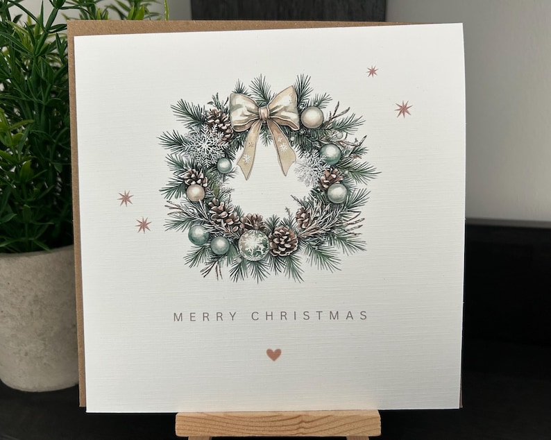 May include: A Christmas card with a watercolour illustration of a wreath. The wreath is composed of pine needles, pine cones, and baubles in green, white, and silver. A bow sits at the top, and the words "MERRY CHRISTMAS" are printed below the wreath.