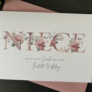 K&ouml;nnte beinhalten: Eine wei&szlig;e Karte mit dem Wort "NIECE" in rosa Buchstaben, die mit rosa Blumen verziert sind. Der Text "With love Sarah on your Thirtieth Birthday" steht unter dem Wort "NIECE".