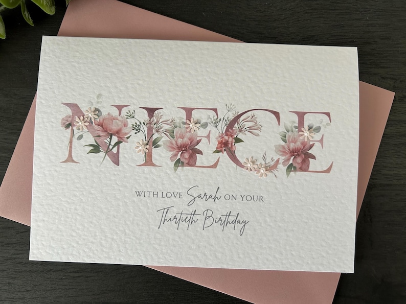 K&ouml;nnte beinhalten: Eine wei&szlig;e Karte mit dem Wort "NIECE" in rosa Buchstaben, die mit rosa Blumen verziert sind. Der Text "With love Sarah on your Thirtieth Birthday" steht unter dem Wort "NIECE".