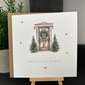 May include: A Christmas card with a watercolour illustration of a wooden door decorated with a wreath, with two small Christmas trees on either side. The words "MERRY CHRISTMAS" are printed below the door, with a small heart symbol.