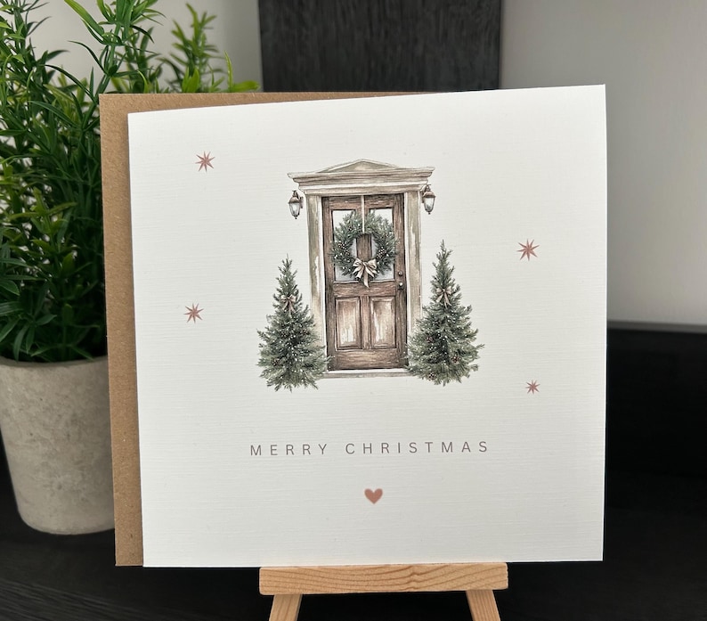 May include: A Christmas card with a watercolour illustration of a wooden door decorated with a wreath, with two small Christmas trees on either side. The words "MERRY CHRISTMAS" are printed below the door, with a small heart symbol.