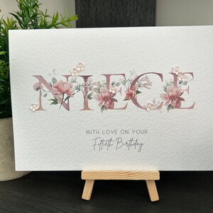 K&ouml;nnte beinhalten: Eine wei&szlig;e Karte mit dem Wort "NIECE" in pinken Blumenbuchstaben. Die Karte tr&auml;gt den Text "With love on your Fiftieth Birthday" unter den Blumenbuchstaben.