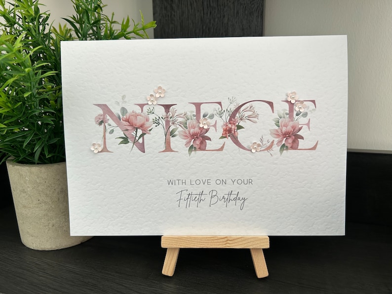 K&ouml;nnte beinhalten: Eine wei&szlig;e Karte mit dem Wort "NIECE" in pinken Blumenbuchstaben. Die Karte tr&auml;gt den Text "With love on your Fiftieth Birthday" unter den Blumenbuchstaben.