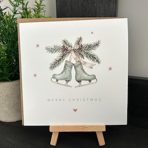 May include: A Christmas card with a watercolour illustration. Two ice skates are shown beneath a sprig of pine and pinecones, tied with a ribbon. The words "MERRY CHRISTMAS" are printed below the skates, with a small heart.