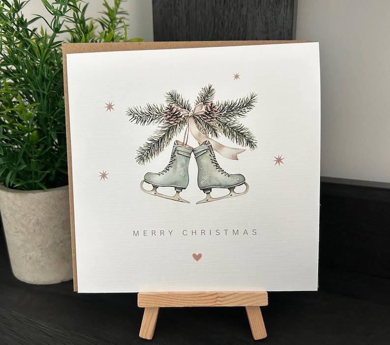 May include: A Christmas card with a watercolour illustration. Two ice skates are shown beneath a sprig of pine and pinecones, tied with a ribbon. The words "MERRY CHRISTMAS" are printed below the skates, with a small heart.