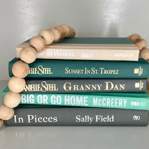 May include: A stack of five books with teal and gray covers. The books are arranged from largest to smallest. The titles of the books are "Op Devotions", "Sunset in St. Tropez", "Granny Dan", "Big or Go Home", and "In Pieces". A wooden bead garland is draped over the top of the stack.