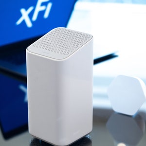 Puede incluir: Un router inal&aacute;mbrico blanco con una parte superior texturizada y un peque&ntilde;o dispositivo hexagonal blanco en el fondo. El router est&aacute; sentado sobre una superficie reflectante. El texto "xFi" es visible en una pantalla azul en el fondo.