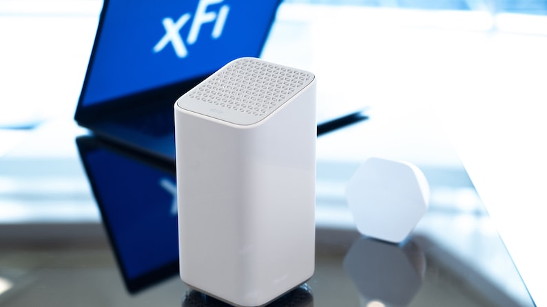 Puede incluir: Un router inal&aacute;mbrico blanco con una parte superior texturizada y un peque&ntilde;o dispositivo hexagonal blanco en el fondo. El router est&aacute; sentado sobre una superficie reflectante. El texto "xFi" es visible en una pantalla azul en el fondo.