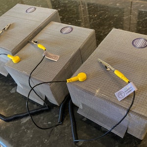 Op de afbeelding: Drie grijze metalen dozen met een cirkelvormig ontwerp op de bovenkant. Elke doos heeft een zwarte draad met een gele sonde en een clip eraan bevestigd. De dozen zijn gelabeld met "CONNECT TO LEARN".