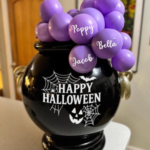 May include: A black cauldron-shaped container with gold handles, filled with purple balloons. The balloons have names written on them. The cauldron has a Halloween-themed design with the words "Happy Halloween".