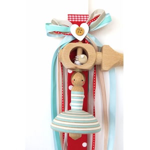 May include: A wooden baby rattle with a blue and white striped doll and a spinning top. The rattle is decorated with red and white gingham ribbon, white polka dot ribbon, and light blue ribbon.