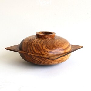 May include: A handcrafted wooden bowl with a lid. The bowl is made of olive wood, featuring a natural wood grain pattern. The lid has a small, cylindrical handle. The bowl sits on a square base, creating a unique design.