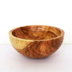 May include: A round wooden bowl made from olive wood. The bowl has a natural wood finish and a light brown color. The grain of the wood is visible and adds to the rustic charm of the bowl.