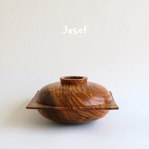 May include: A handcrafted wooden bowl with a lid. The bowl is made of a light brown wood with a unique grain pattern. The lid has a small, cylindrical handle. The word "Josef" is printed in white on the top.