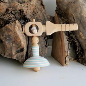 May include: A wooden spinning top toy featuring a figure with a painted face and a light blue and white striped skirt. The toy includes a separate wooden handle mechanism with a white string for winding and launching the spinning top.